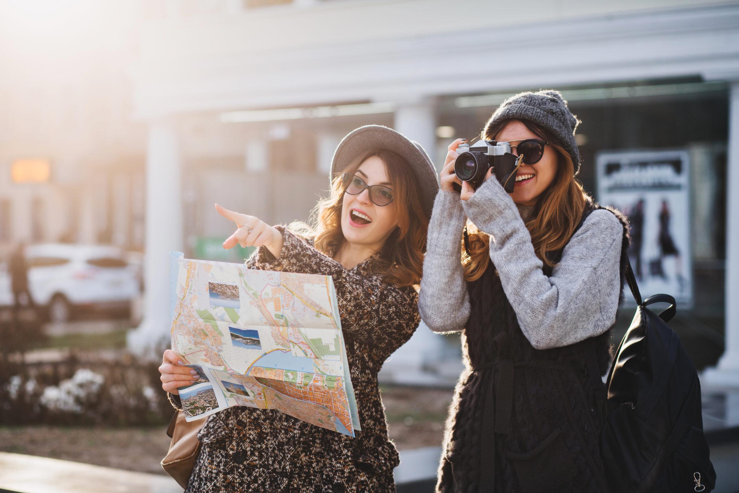 happy travel together of two fashionable girls in sunny city centre. young joyful women expressing positivity, using map, vacation with bags, camera, making photo, cheerful emotions, great mood.